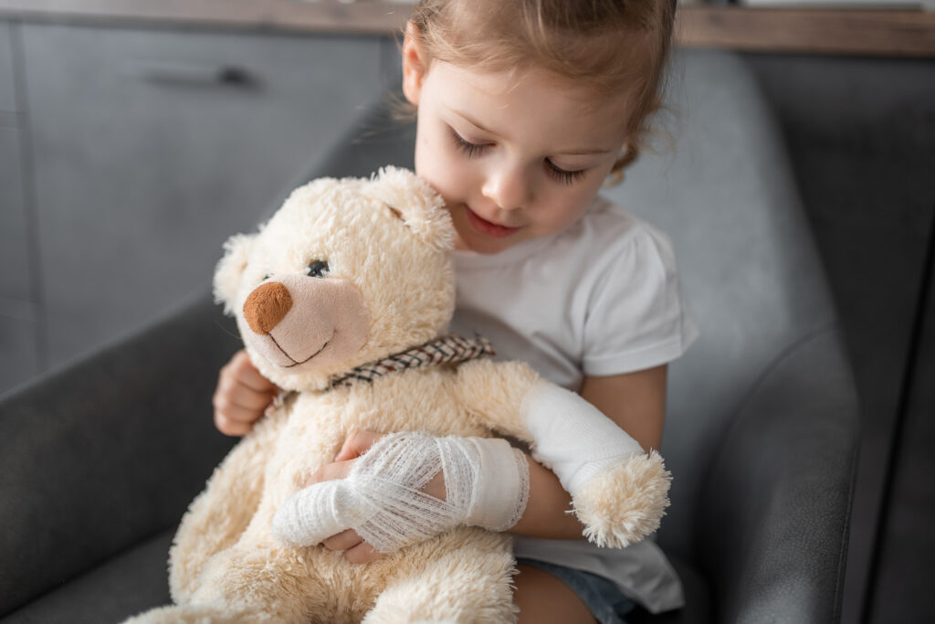 Little girl with broken finger holds teddy bear with a bandaged paw at the doctor's appointment in